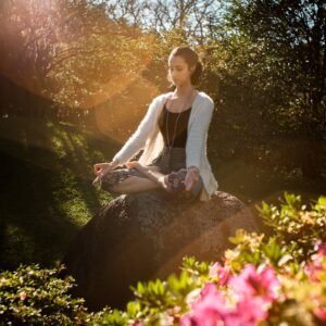 Woman in tranquil meditation pose outdoors, bathed in sunlight, surrounded by lush greenery.